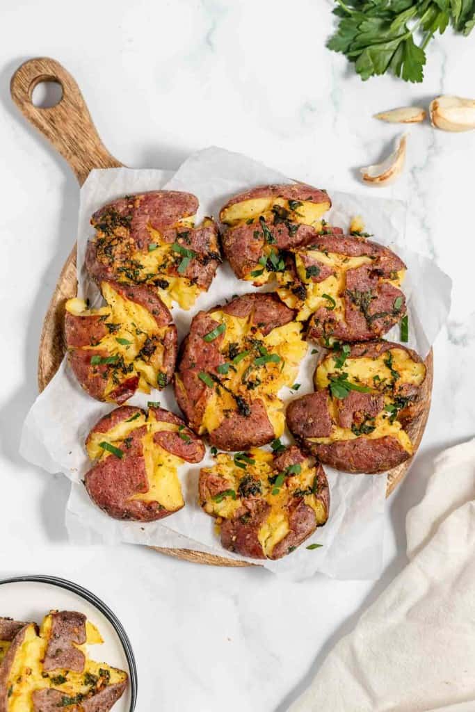 A wooden tray with several smashed potatoes garnished with herbs and placed on parchment paper. Garlic cloves and a sprig of parsley are placed nearby on a white surface.