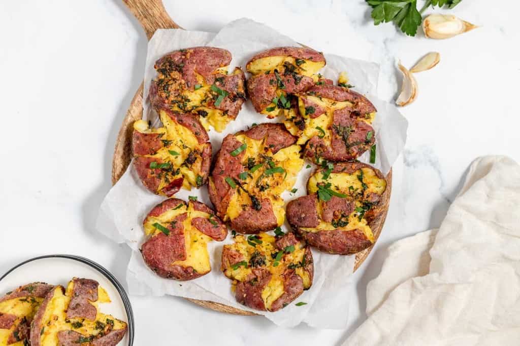 A wooden tray with several smashed potatoes garnished with herbs and placed on parchment paper. Garlic cloves and a sprig of parsley are placed nearby on a white surface.
