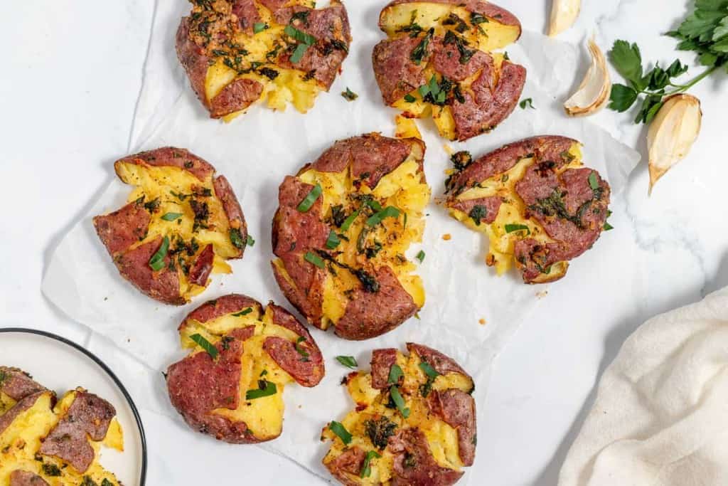 Crispy smashed potatoes topped with fresh herbs and seasoning, arranged on a white parchment paper with garlic cloves and parsley on the side.