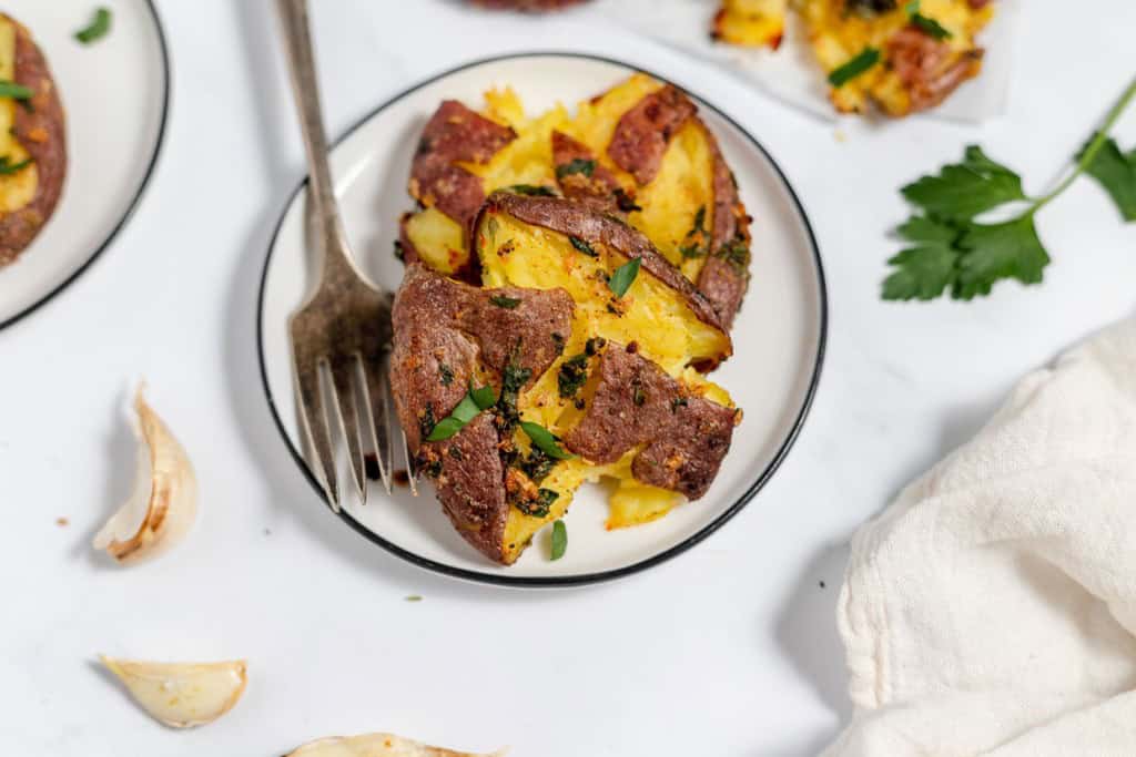 A small white plate with smashed and roasted potatoes garnished with chopped parsley. A fork is placed on the plate's edge. Garlic cloves and a parsley leaf are visible on the table.