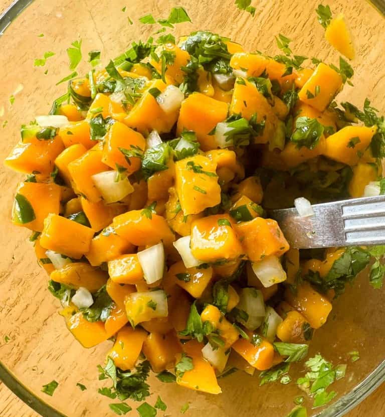 A glass bowl contains a fresh mango salsa with diced mangoes, onions, and chopped cilantro, mixed with a fork.