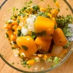 Close-up of a glass bowl containing a mango salsa mix with diced mango, onions, cilantro, and a fork lifting some of the mixture.