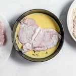 A piece of floured meat is being dipped into an egg mixture, with more floured meat on a plate to the left and a bowl of seasoned flour to the right.