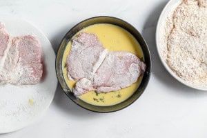 A piece of floured meat is being dipped into an egg mixture, with more floured meat on a plate to the left and a bowl of seasoned flour to the right.