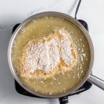 Chicken breast coated in flour frying in hot oil inside a stainless steel pan on a stovetop.