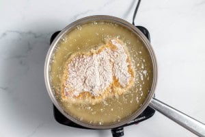 Chicken breast coated in flour frying in hot oil inside a stainless steel pan on a stovetop.