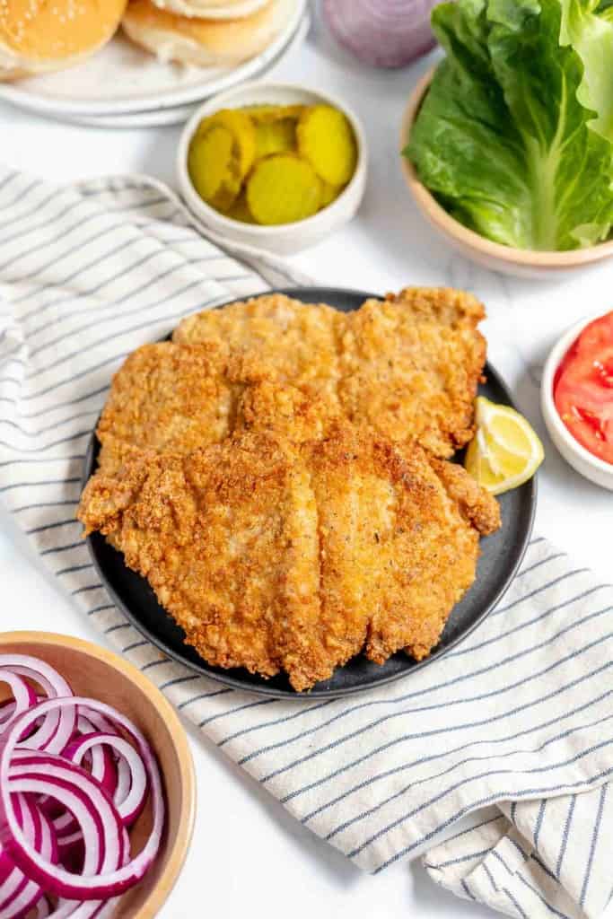Three breaded and fried schnitzels on a black plate, garnished with a lemon wedge, surrounded by sliced pickles, lettuce, tomato, and onion on a striped cloth.