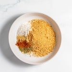 A white bowl contains breadcrumbs, shredded coconut, and various ground spices, placed on a white marble surface.