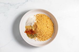 A white bowl contains breadcrumbs, shredded coconut, and various ground spices, placed on a white marble surface.