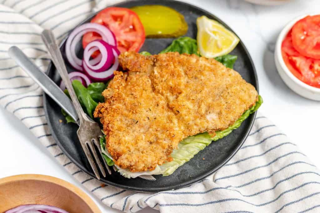 A plate with a crispy breaded fried chicken cutlet, lettuce, tomato slices, red onion rings, pickle, and lemon wedge. Utensils are placed on the plate. A stripped cloth is underneath.