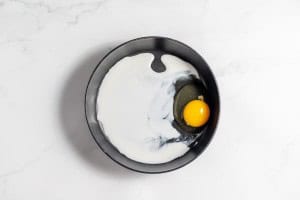 A black bowl containing a raw egg yolk and a splash of milk rests on a white marble surface.