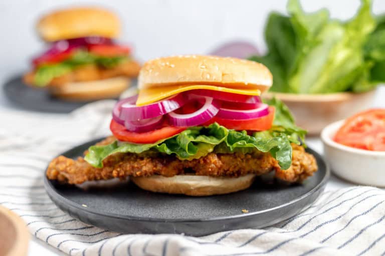 A fried chicken sandwich with lettuce, tomato, red onion, and cheese sits on a black plate. Another sandwich and fresh ingredients are blurred in the background.
