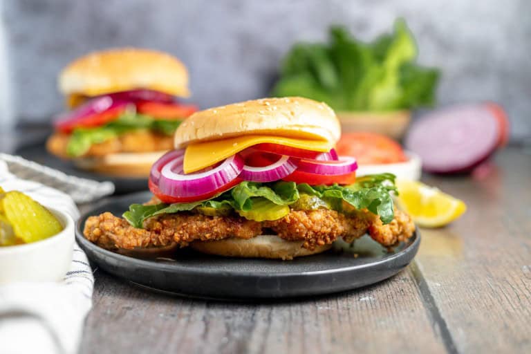 A close-up of a fried chicken sandwich with lettuce, red onions, and cheese on a plate. A second sandwich, pickles, lettuce, and sliced red onions are in the background.
