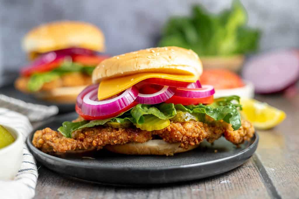 A crispy chicken sandwich with lettuce, red onion, and cheese on a sesame seed bun, served on a black plate. Another sandwich and vegetables are blurred in the background.