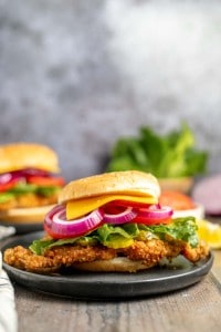 A close-up of a crispy chicken sandwich with lettuce, red onions, and cheddar cheese on a sesame seed bun, placed on a black plate. Another sandwich and some vegetables are blurred in the background.