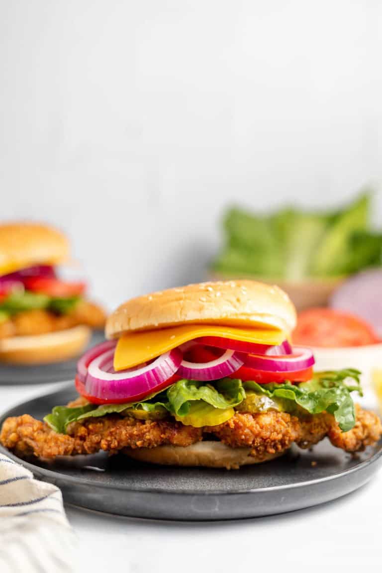 A crispy chicken sandwich with lettuce, tomato, cheese, and red onion on a sesame seed bun, served on a grey plate. Another sandwich and ingredients are blurred in the background.