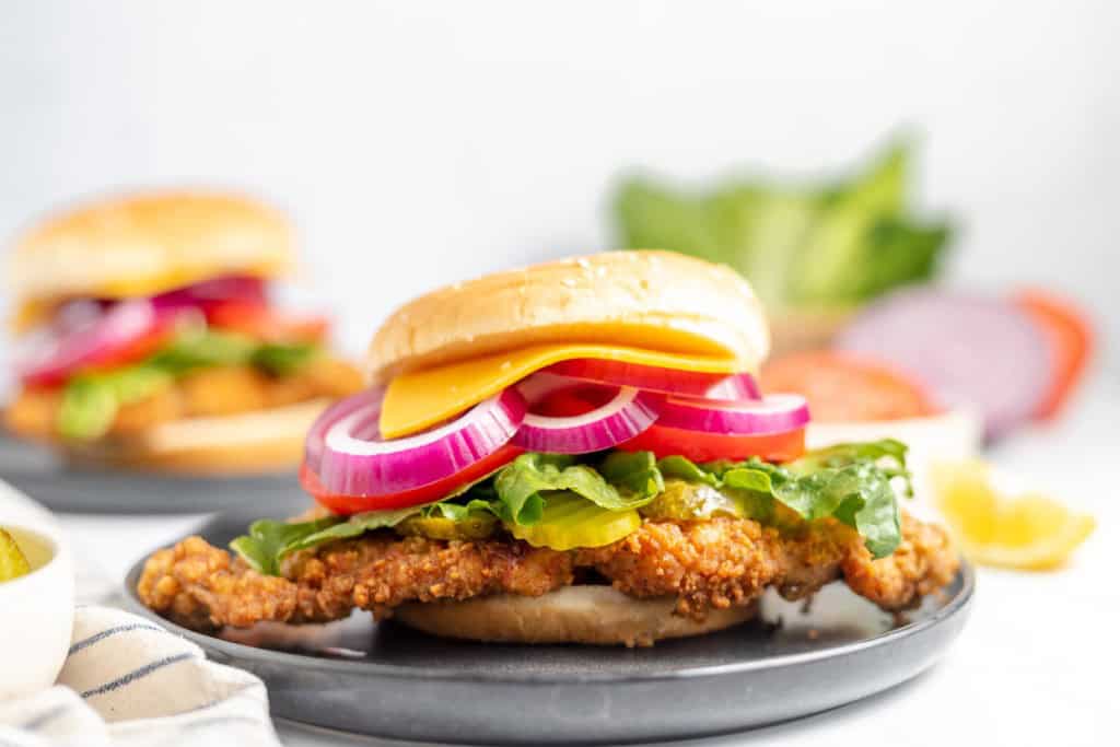 A plate holding a fried chicken sandwich topped with lettuce, pickles, red onion, cheese, and a bun, with another sandwich blurred in the background.