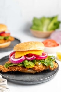 A crispy chicken sandwich with cheese, red onion slices, and lettuce on a sesame seed bun, served on a black plate. Additional ingredients are visible in the background.