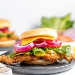 A close-up of a fried chicken sandwich topped with lettuce, red onion, and cheese on a plate. Another sandwich and sliced vegetables are in the blurred background.
