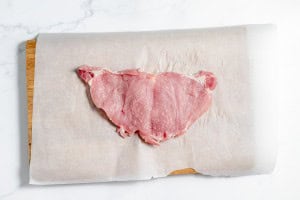 A piece of raw pork schnitzel laid on parchment paper over a wooden surface.