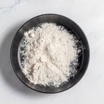 A black bowl filled with white flour sits on a white marble surface.