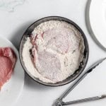 Raw breaded cutlet in a black bowl of flour, with a piece of raw meat nearby on a white plate, accompanied by metal tongs.