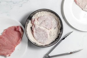 Raw breaded cutlet in a black bowl of flour, with a piece of raw meat nearby on a white plate, accompanied by metal tongs.