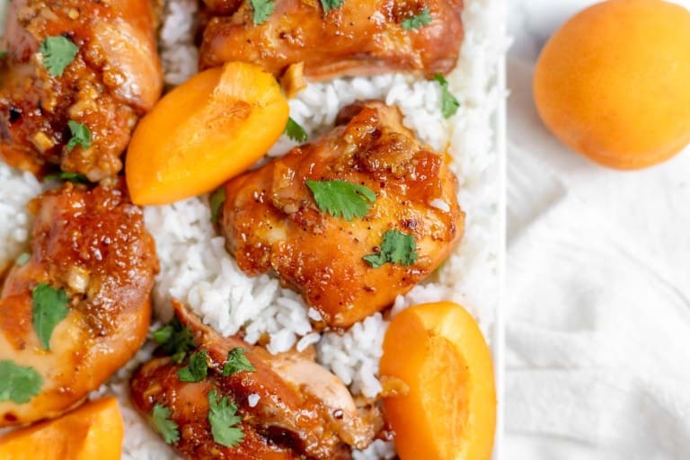 Close-up of roasted chicken pieces garnished with cilantro, placed on a bed of white rice with fresh apricot slices.