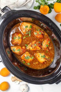 Slow cooker filled with cooked chicken thighs in an orange sauce, garnished with fresh herbs, with apricots and cilantro on the side.