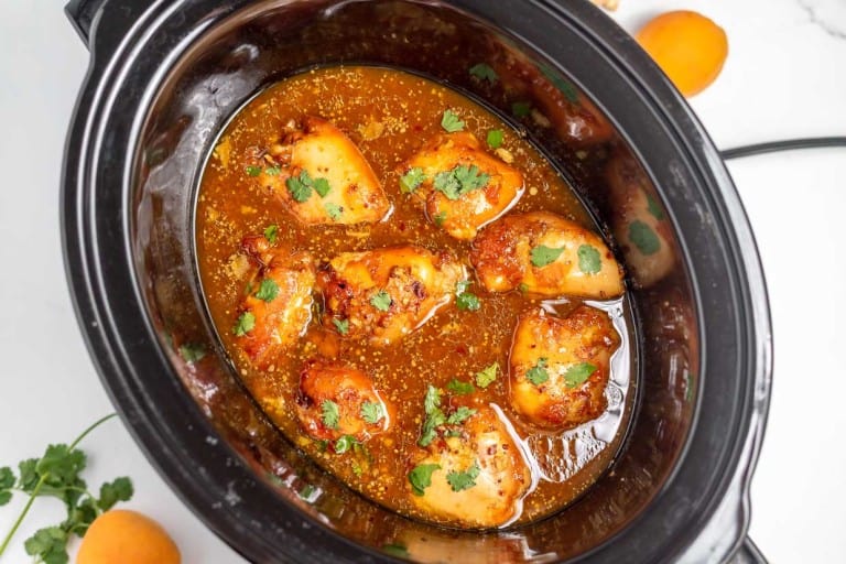 Image of a slow cooker containing cooked chicken thighs in a savory sauce, garnished with fresh cilantro. Orange slices and cilantro leaves are visible beside the slow cooker.