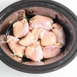 Raw chicken thighs placed inside a black slow cooker on a white surface.
