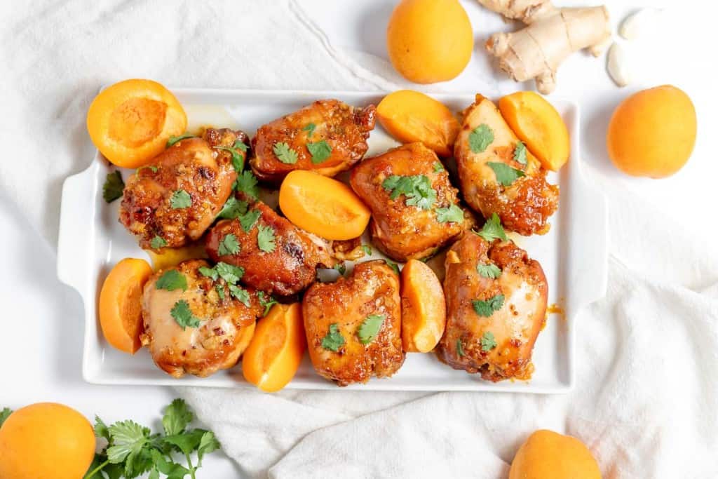 A white rectangular platter with cooked chicken thighs garnished with cilantro and halved apricots, surrounded by whole apricots and ginger pieces on a white cloth.