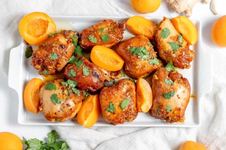 A rectangular white plate filled with cooked chicken thighs garnished with fresh cilantro and surrounded by halved apricots, placed on a white tablecloth.