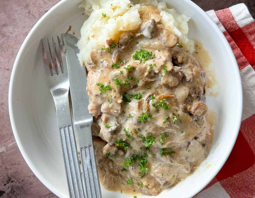 A bowl of mashed potatoes topped with a creamy mushroom sauce, resembling those comforting slow cooker recipes, with a fork and knife placed on the side.