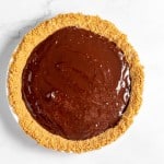 A chocolate pie with a graham cracker crust is displayed on a white marble surface. The filling is smooth and glossy.