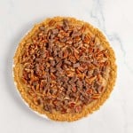 A pie with a crumbly crust, topped with chopped pecans and pieces of chocolate, displayed on a white marble surface.