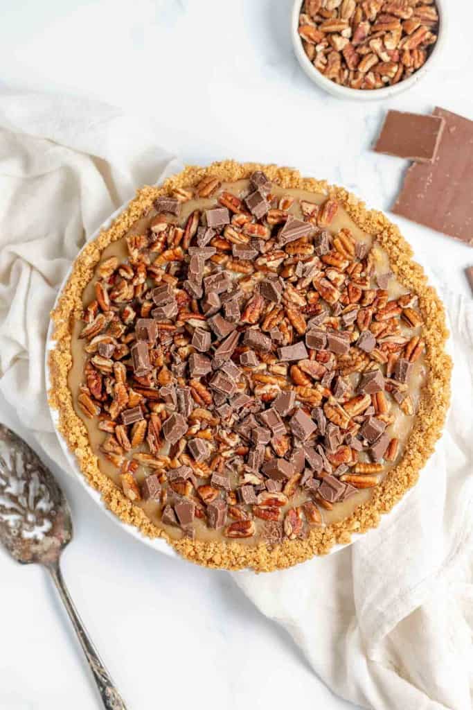 A sliced pie with a graham cracker crust, topped with chopped pecans and chocolate chunks, placed on a white cloth. A bowl of pecans and pieces of chocolate are next to the pie.
