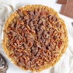 A sliced pie with a graham cracker crust, topped with chopped pecans and chocolate chunks, placed on a white cloth. A bowl of pecans and pieces of chocolate are next to the pie.