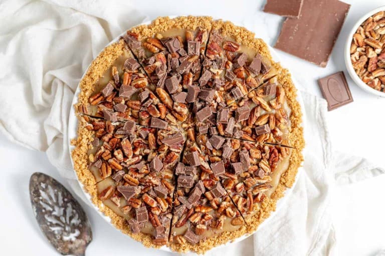 A sliced pie with a graham cracker crust, topped with chopped pecans and chocolate chunks, placed on a white cloth. A bowl of pecans and pieces of chocolate are next to the pie.