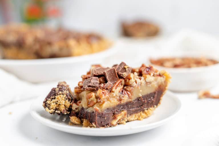 A close-up of a slice of layered dessert with a graham cracker crust, chocolate layer, and pecan topping on a white plate. Other desserts are blurred in the background.
