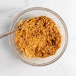 A glass bowl filled with crumbled graham cracker mixture with a spoon on a white marble surface.