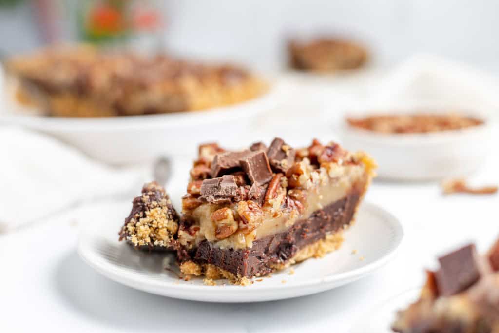 A slice of pie with chocolate chunks and pecans sits on a white plate. A fork is resting on the plate, and a whole pie is visible in the background.