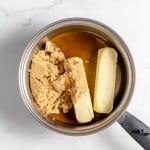 A saucepan on a marble surface containing two sticks of butter, brown sugar, and a liquid mixture.
