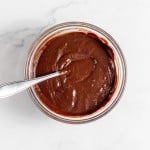 A glass bowl filled with chocolate ganache, with a metal spoon partially submerged in the ganache, sitting on a white marble surface.
