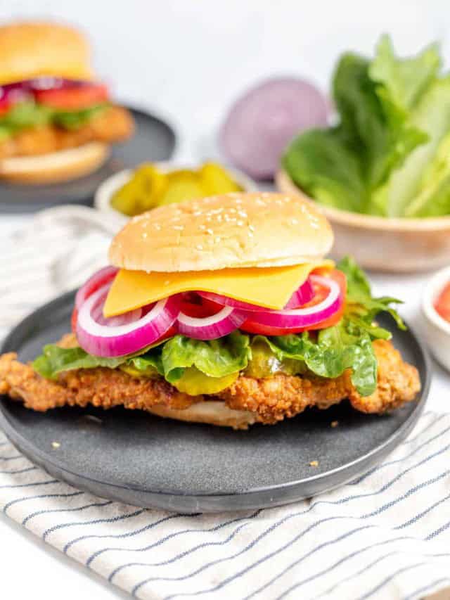 A fried chicken sandwich with lettuce, pickles, red onion, and a slice of cheese on a sesame seed bun, placed on a black plate with an additional sandwich, lettuce, and onion in the background.