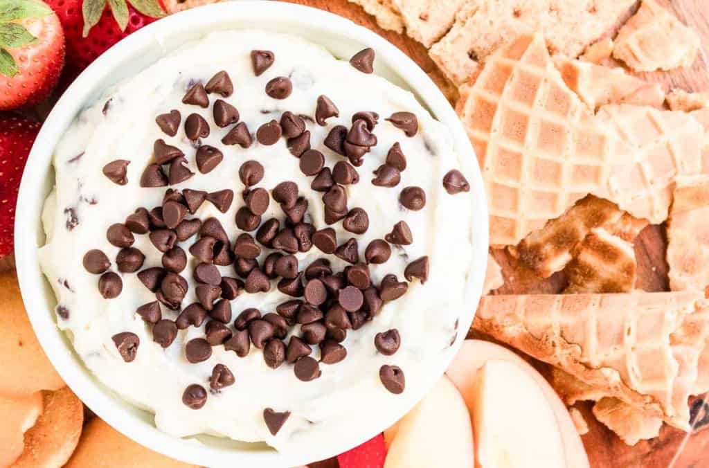 A bowl of creamy dessert dip topped with chocolate chips, surrounded by waffle pieces, apple slices, and strawberries.