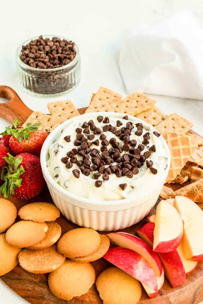 A platter with a bowl of chocolate chip dip surrounded by vanilla wafers, apple slices, strawberries, graham crackers, and waffle pieces. A small jar of chocolate chips is in the background.