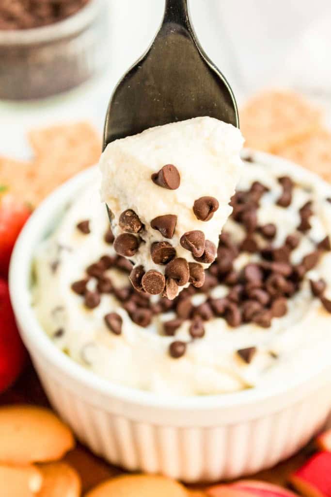 A spoon holding a portion of cream cheese with chocolate chips above a white bowl filled with the same mixture, surrounded by strawberries and bread pieces.