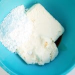 A blue mixing bowl containing cream cheese, powdered sugar, and vanilla extract on a white surface.