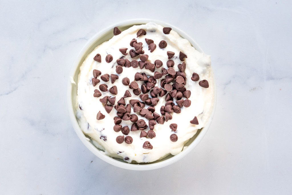 A bowl of white creamy dip topped with chocolate chips on a white marble surface.